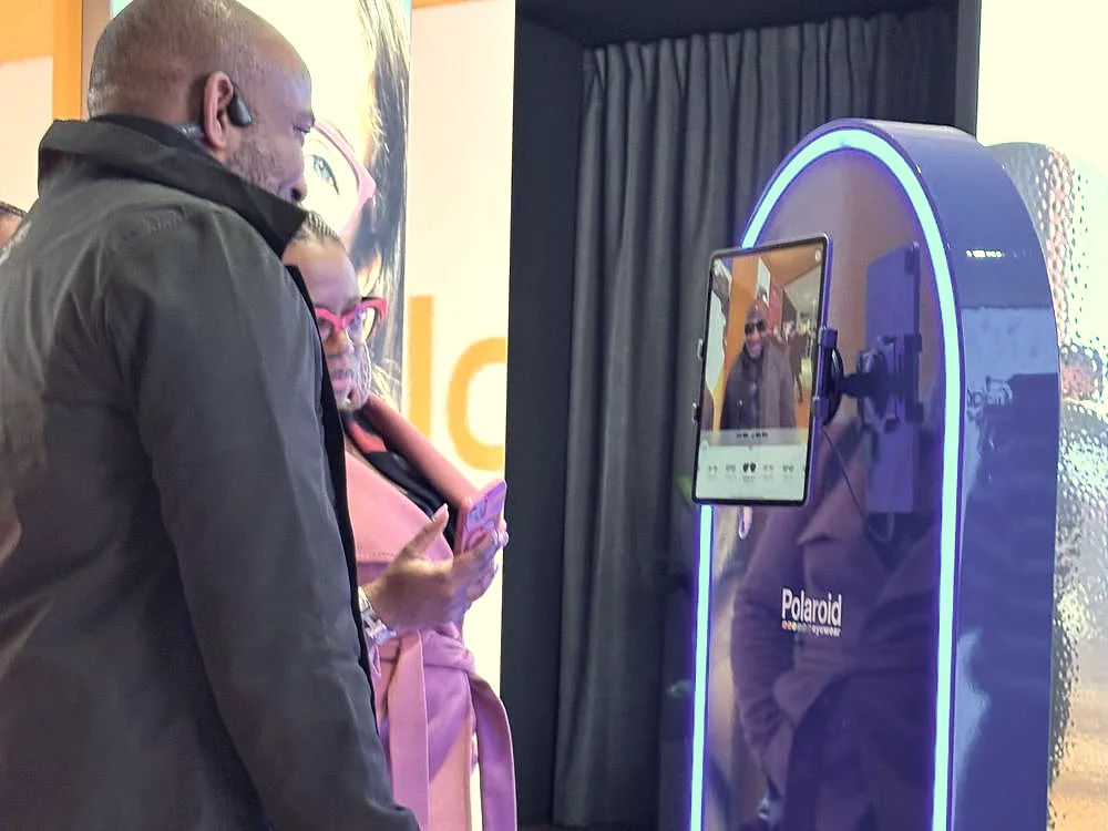In a Polaroid space, a visitor looks at himself on the installation's tablet while his partner checks her phone.