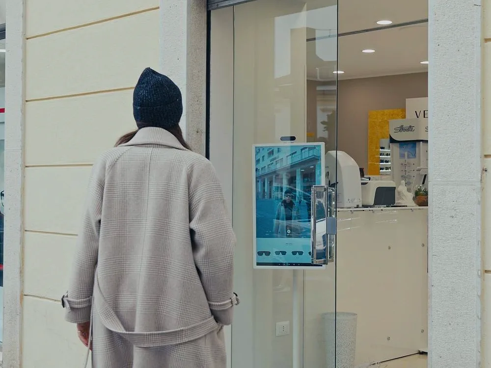 A passer-by stops in front of an optician's shop window, where a digital mirror catches her in frame from the street.