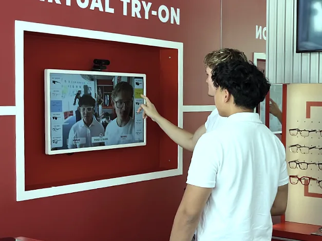 Two young people in front of a screen set into a red wall with the words Virtual Try-On: they see themselves together, each wearing their own glasses.
