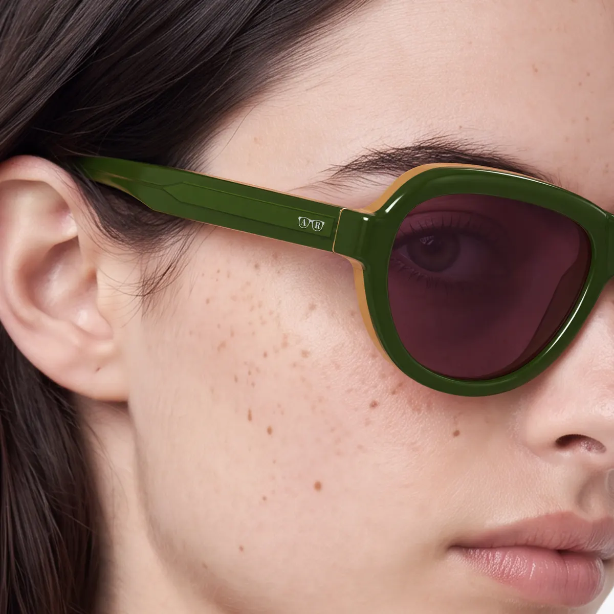 Primo piano di un occhiale da sole Rover indossato da una donna con capelli scuri: acetato verde oliva con riflessi visibili, lente marrone e piccolo logo in metallo sull'asta.